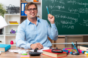 happy-smiling-young-male-teacher-sitting-school-desk-with-books-notes-holding-pencil-front-blackboard-classroom